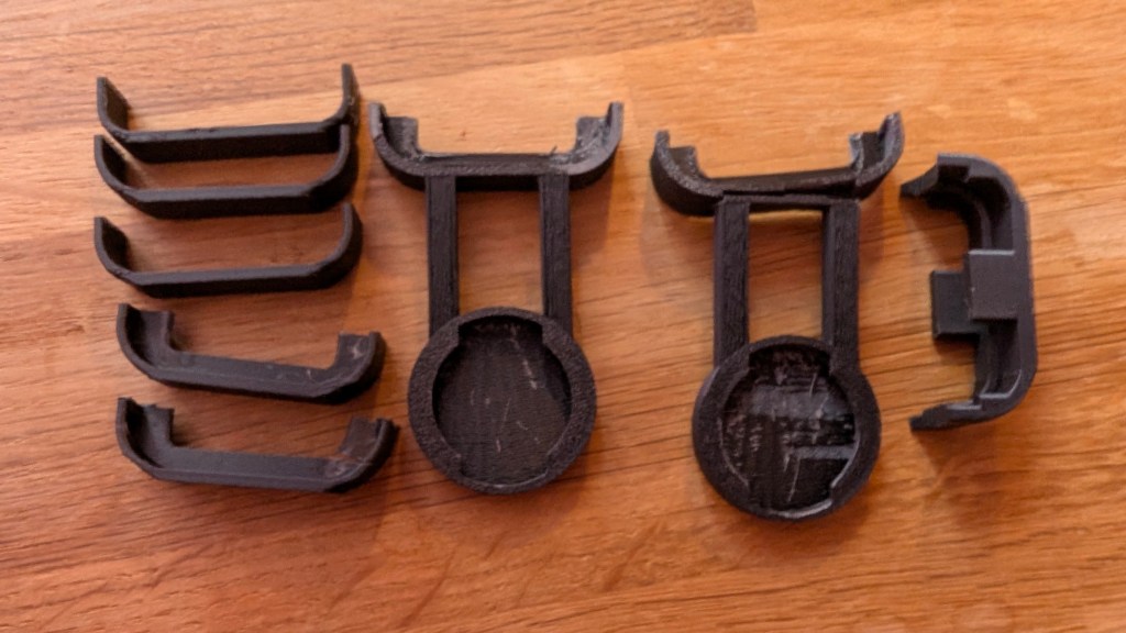 A series of incomplete or broken 3D printed Garmin bike mount prototypes, laid out on a wooden table. The final piece on the right is visibly snapped.