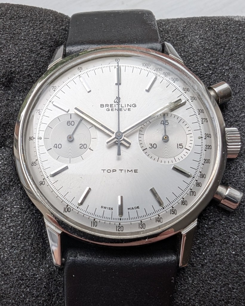 Close-up of a vintage silver Breitling Top Time watch with a black suede strap, featuring a minimalist dial with two subdials and sleek silver hands.