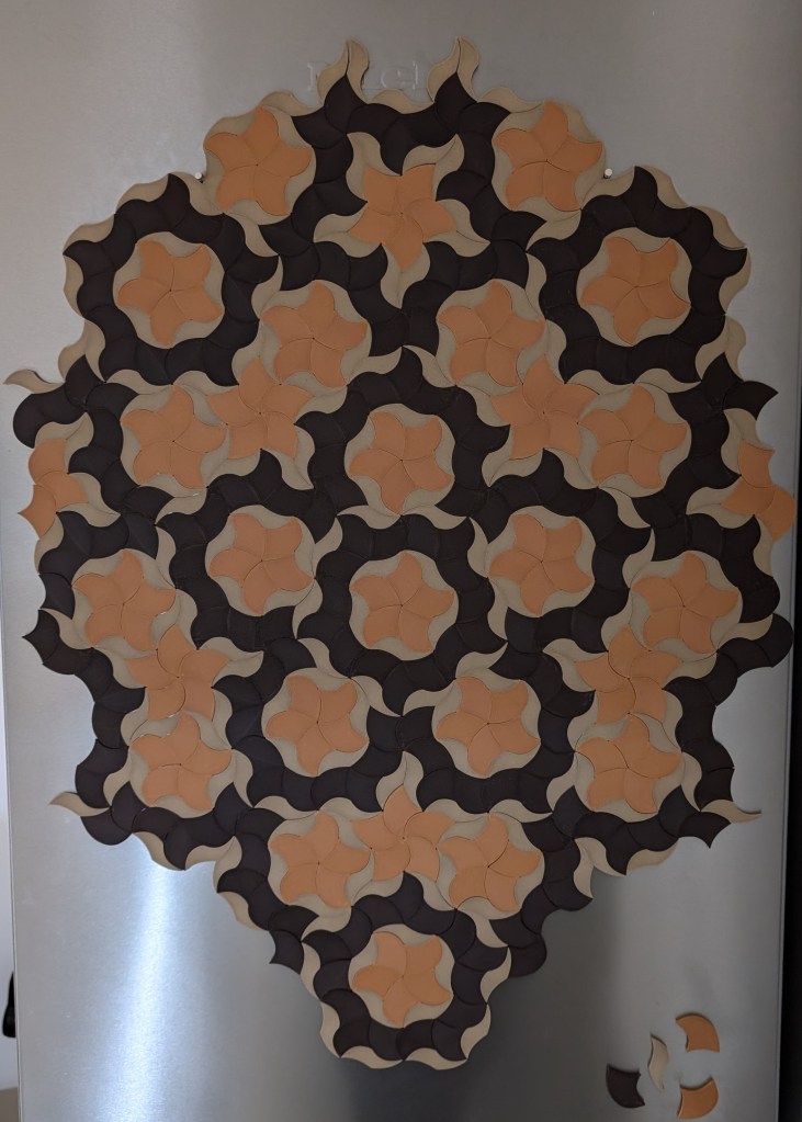 A fridge covered in an intricate Penrose tiling pattern made from interlocking 3D-printed magnets in orange, black, and beige. The tiling forms a mesmerizing, non-repeating pattern—except for the bottom corner, where a few pieces remain unfinished, hinting at the obsessive journey behind it.