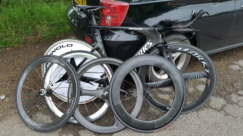 5 different time trial front wheels, including deep-section and lightweight wheels, leaned against a car beside a time trial bike, suggesting excessive preparation and equipment choices.
