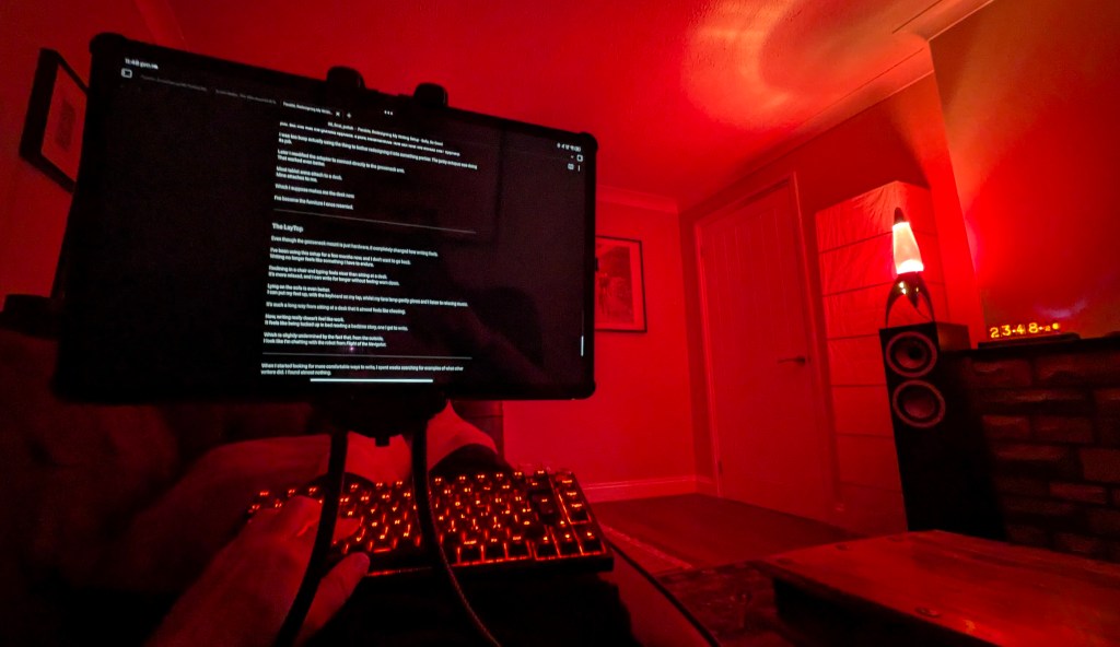 A first-person perspective of a cozy, low-light living room bathed in a deep red glow from a rocket-shaped lava lamp sitting on a tall bookshelf speaker to the right. In the foreground, a large digital tablet screen displaying markdown text is suspended by a flexible mount directly over a mechanical keyboard with glowing red backlighting, resting on a lap.