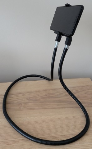 A flexible black gooseneck phone holder rests on a light wood-grained tabletop. It consists of a large, circular loop meant to drape around a person's neck or body, with two parallel flexible arms extending upward to hold a smartphone clamped in landscape orientation against a plain white background.