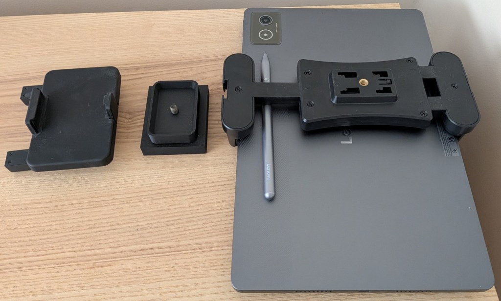 An exploded view of DIY tablet mount components laid out on a wooden desk next to a gray Lenovo tablet. A custom 3D-printed black plastic block with a central screw thread sits between the original smaller device clamp and a much wider spring-loaded tablet holder.