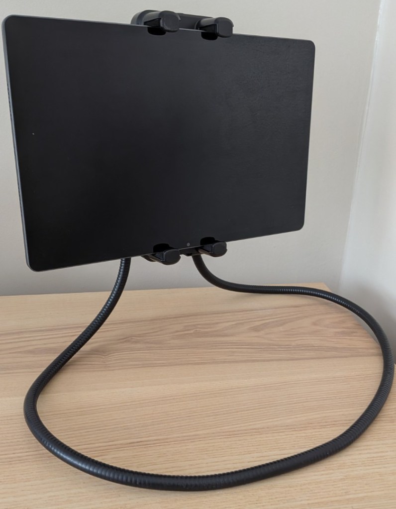 Front three-quarter view of a large black tablet screen securely held by thick plastic brackets, attached to a wide, circular black flexible gooseneck base that rests on a light wood desk.