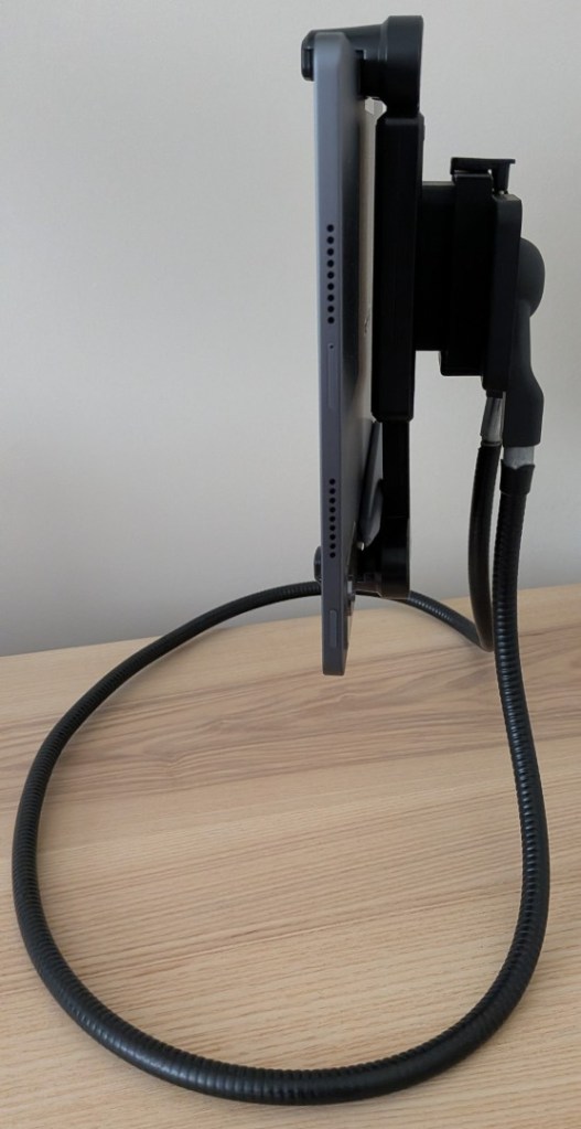 Side profile view of a large tablet clamped into a bulky, multi-layered black plastic mounting bracket assembly. The entire heavy structure is supported by a thick black flexible gooseneck arm resting on a wooden tabletop.
