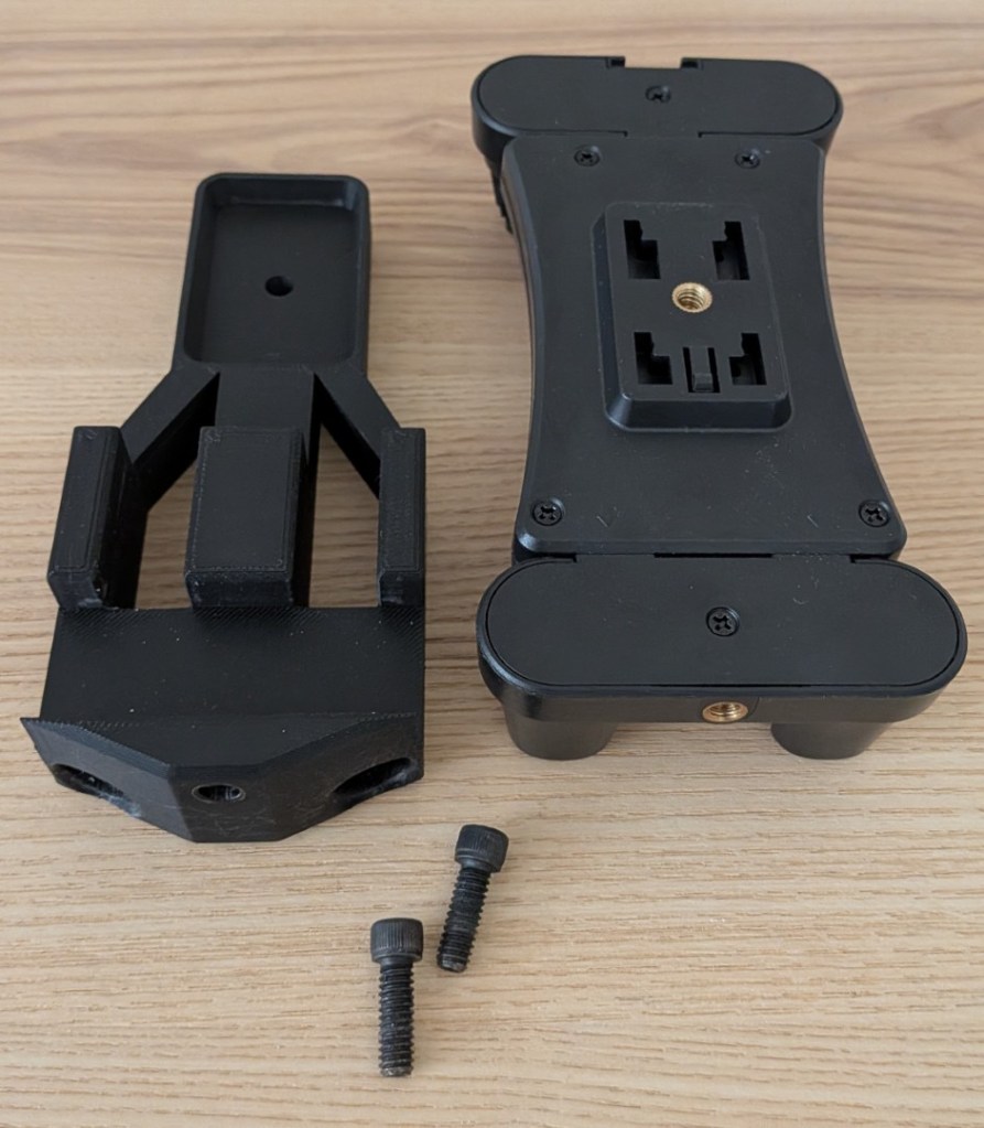 An exploded view of the upgraded DIY mount components on a wood desk. A redesigned black 3D-printed adapter plate featuring two entry holes for a gooseneck frame sits next to a tablet clamp and two black hex bolts.