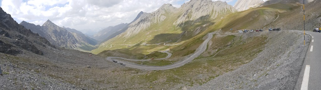 A wide, sweeping panoramic view looking down a vast, desolate alpine valley from a high mountain road. A narrow, paved road with tight switchback curves winds down the steep, rocky mountain slopes. The landscape is stark and primeval, dominated by gray shale, loose rocks, and sparse patches of green grass, with jagged peaks stretching into the hazy distance under a bright, cloudy sky.