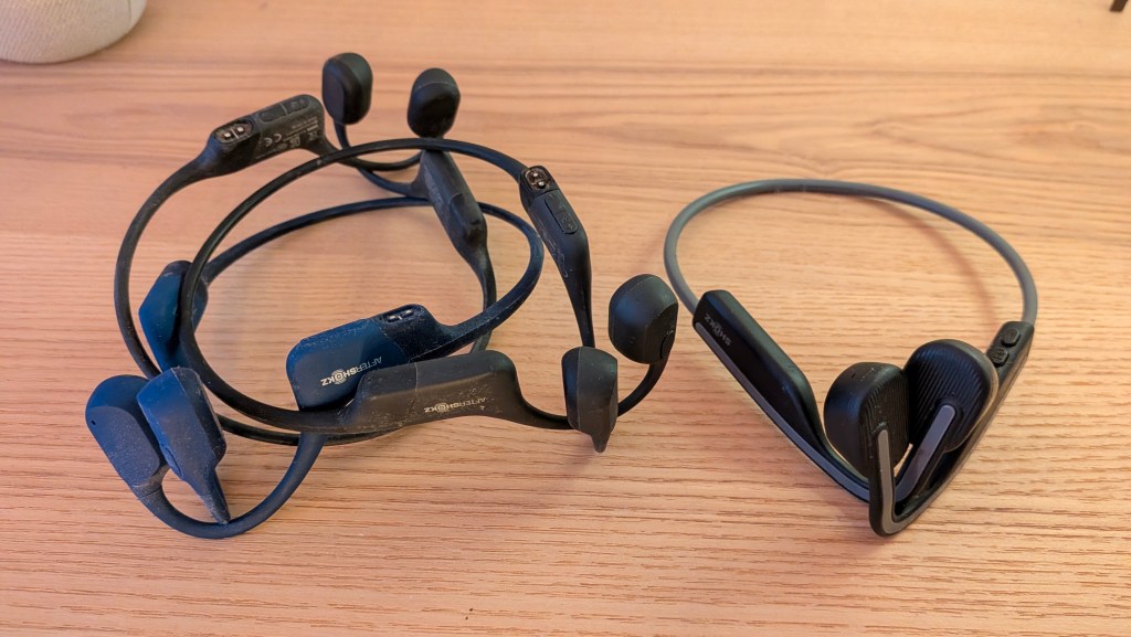 A top-down view on a light wood tabletop showcasing the aftermath of a tech war of attrition. On the left sits a tangled, slightly dusty heap of multiple broken AfterShokz Aeropex bone-conducting headphones. On the right sits a single, clean, and perfectly intact pair of Shokz OpenMove headphones.